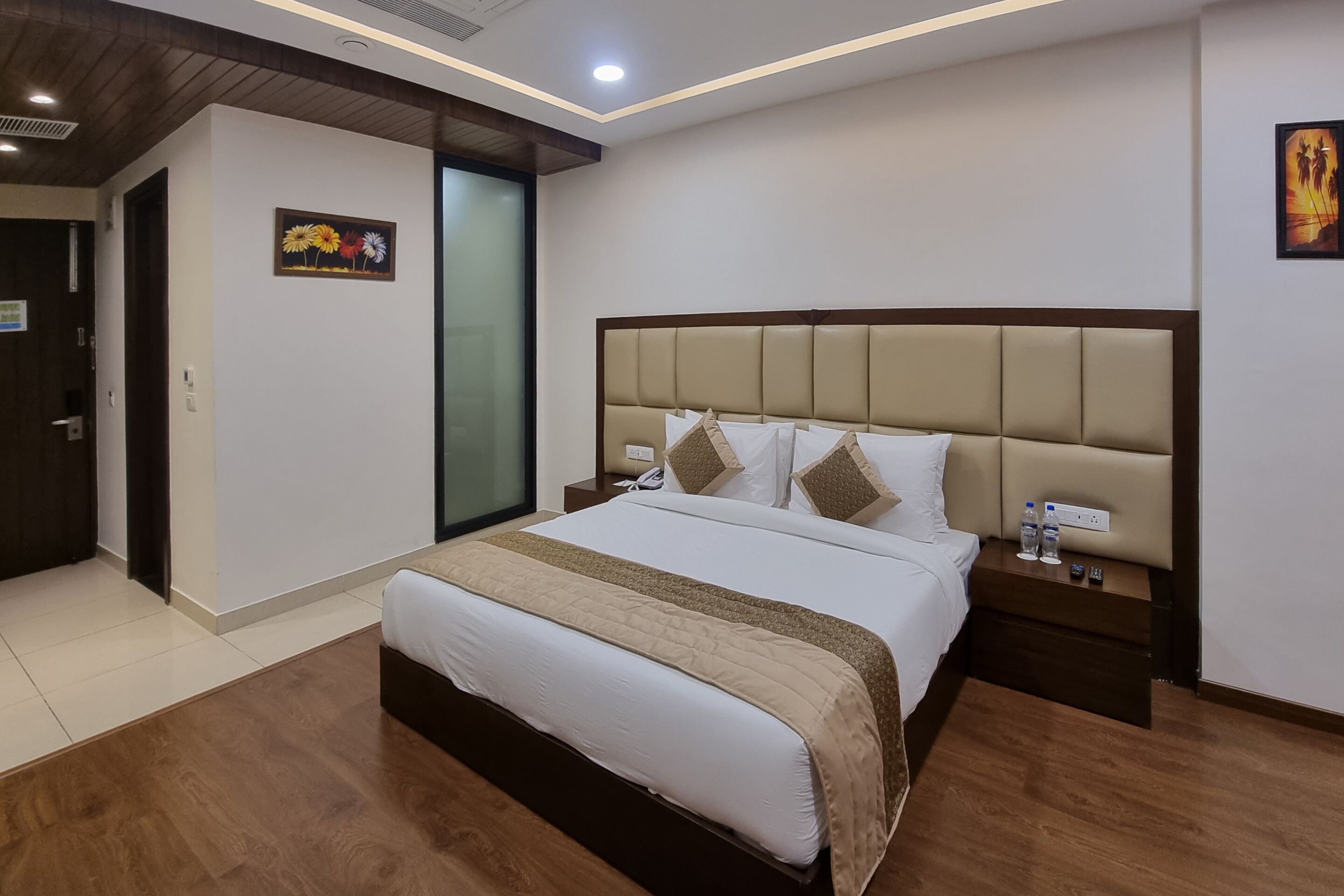 Een hotelkamer met een groot bed, houten vloeren, een nachtkastje met een telefoon en waterflessen, en een schilderij aan de muur.