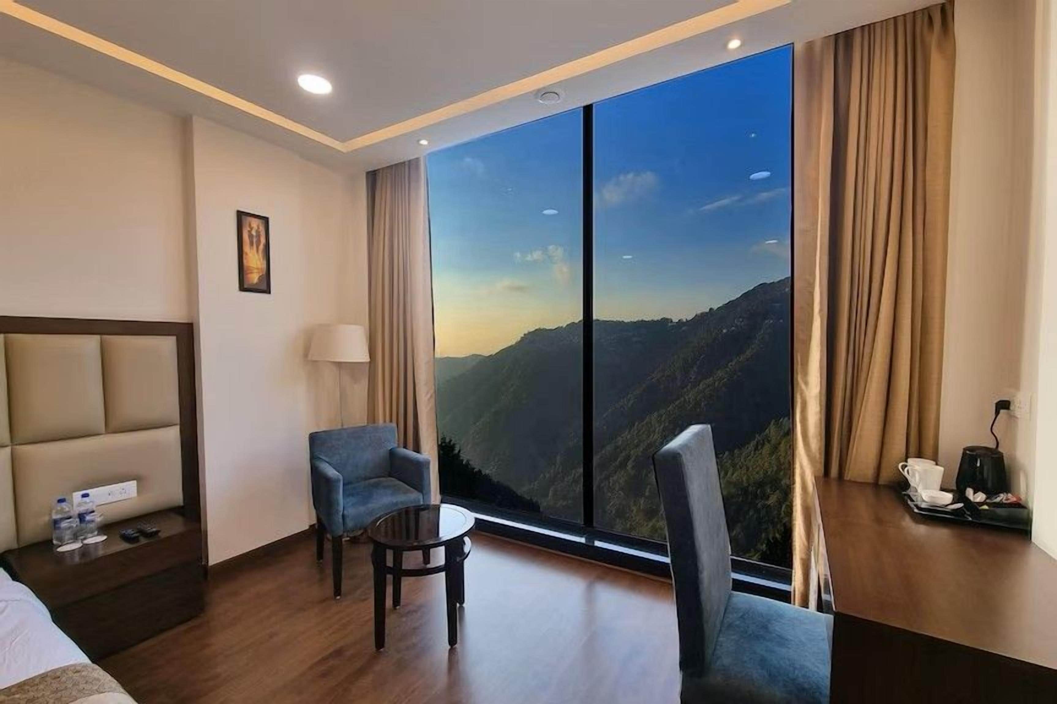 A hotel room with a large window offering a mountain view, a bed, a desk with a chair, and a small table.