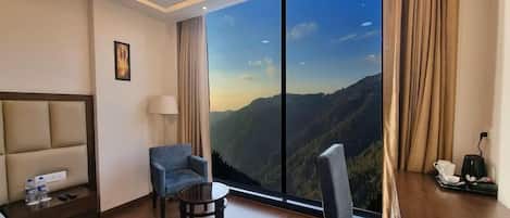 Habitación de hotel con amplia ventana que ofrece vistas a la montaña, cama, escritorio con silla y una mesita.