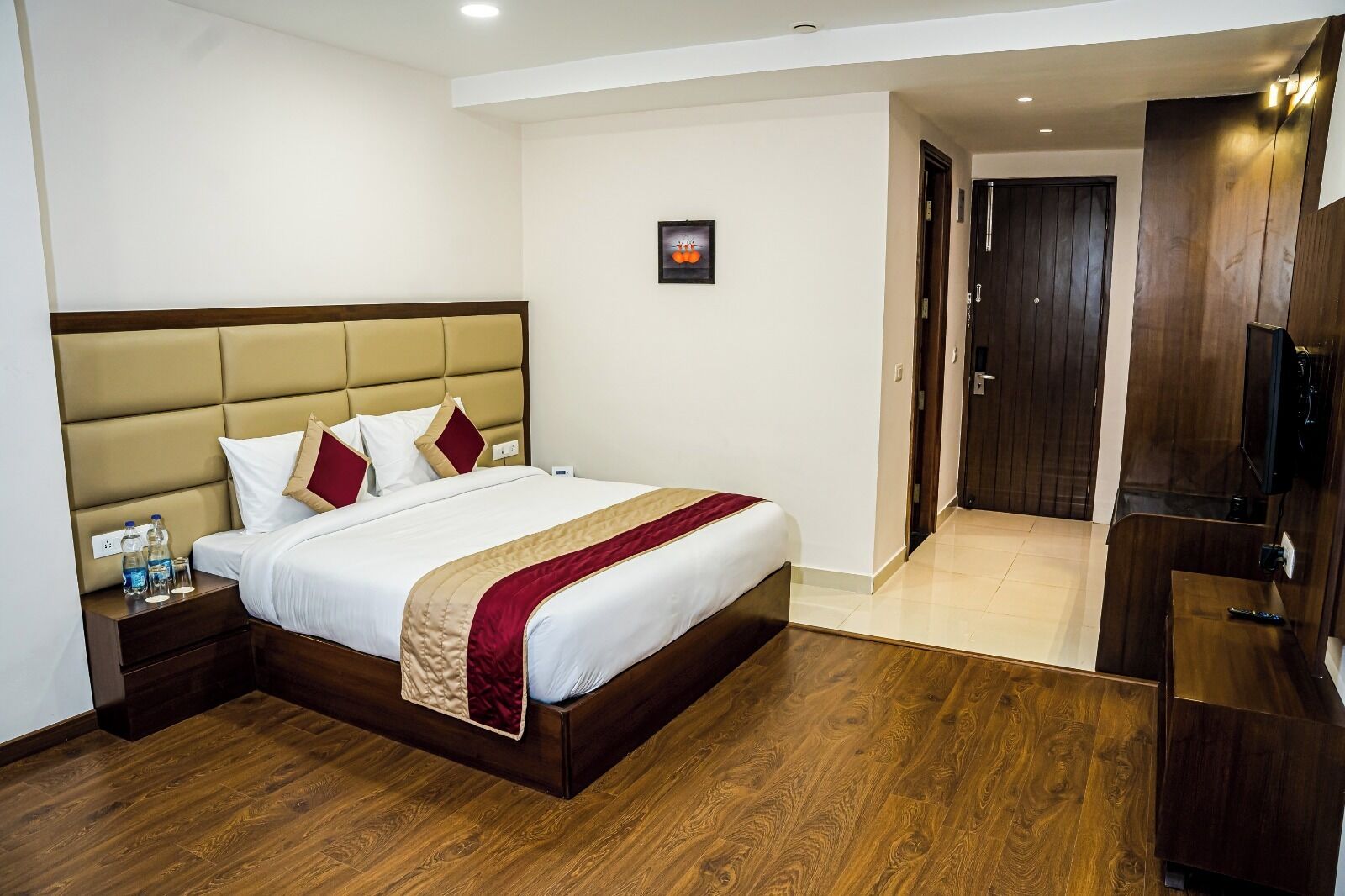 Een hotelkamer met een groot bed, houten vloeren, een hoofdbord, een nachtkastje met waterflesjes en een tv op een houten standaard.