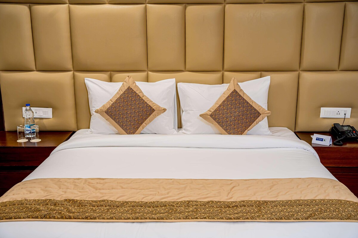 Une chambre d’hôtel avec un grand lit, deux coussins décoratifs, une table de chevet avec une bouteille d’eau, un téléphone et une prise électrique.