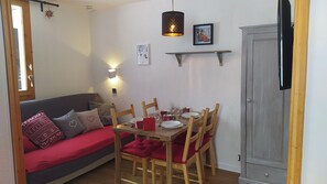 Un espacio de sala acogedor con una mesa de comedor de madera dispuesta para cuatro personas, un sofá gris con cojines rojos, un aparador de madera y un estante fijado a la pared con objetos decorativos.