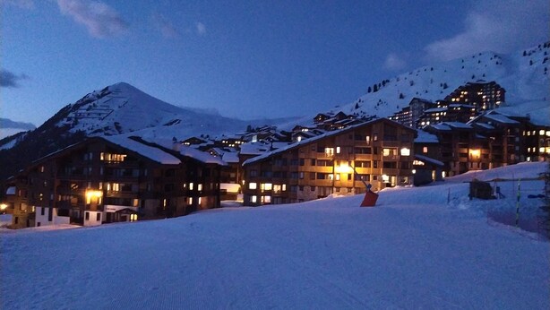 Un complejo turístico en la montaña cubierto de nieve, con varias edificaciones de madera, ventanas iluminadas y una pista de esquí al frente.