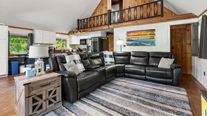 A spacious living area with a large black leather sectional sofa, a wooden dining table, and a kitchen in the background. - Lakefront Home-Stunning Views-Hot Tub, 3100 sqft! (Wakefield)