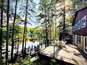 A wooden deck with a view of a lake and forest, a covered outdoor seating area, and a building with large windows. - Lakefront Home-Stunning Views-Hot Tub, 3100 sqft! (Wakefield)