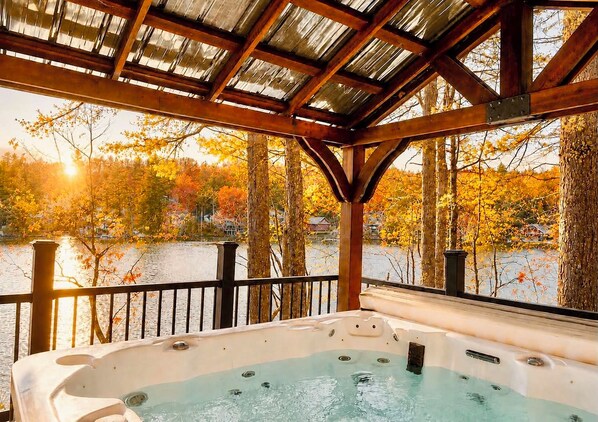 A hot tub under a wooden pergola with a view of a lake and autumn trees.