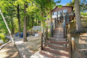 A wooden staircase leading to a cabin with a deck, surrounded by trees and a pond.