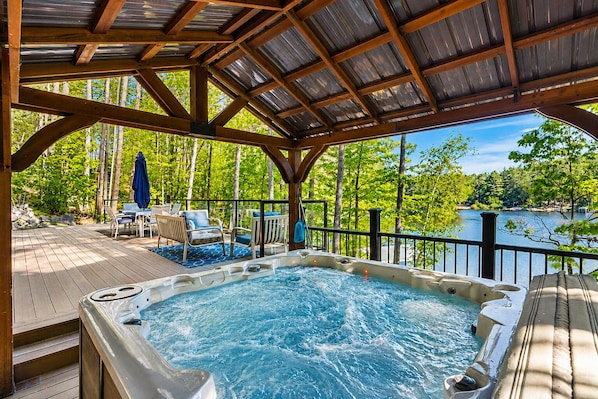 A hot tub with a wooden deck and outdoor furniture, overlooking a lake with trees.