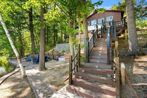 A wooden cabin with a staircase leading to a deck, surrounded by trees and a small pond.