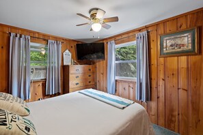 A room with wooden paneling, a bed with a white bedspread, a ceiling fan, a television, and a window with curtains.