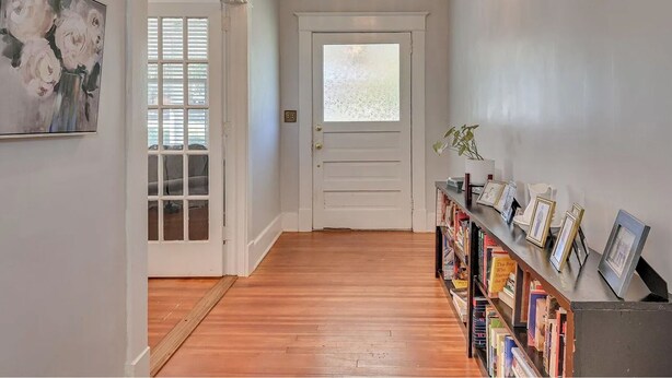 A hallway with a wooden floor, a white door with a frosted glass window, a chandelier, and a bookshelf with framed pictures.