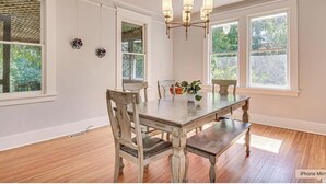 A dining room with a wooden table and chairs, a chandelier, and large windows with a view of greenery.