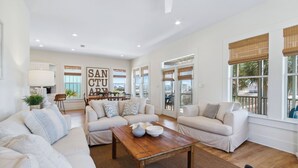 A spacious living room with a white sofa set, a wooden coffee table, and large windows with blinds.