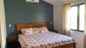A bedroom with a wooden bed, floral bedding, a nightstand, and a window with curtains. - 2 Bed 2 Bath condo with pool, full kitchen, in the marine sunset condos. (Playa del coco)