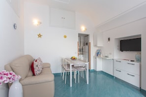 A compact living space with a kitchenette, a dining area with a table and chairs, a sofa with cushions, and a TV mounted on the wall.
