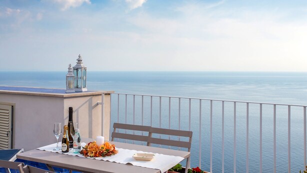 Une table dressée avec des verres à vin, des bouteilles et une bougie, sur un balcon donnant sur la mer.