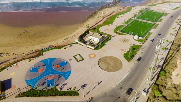 An aerial view of a coastal area with a beach, a sports court, and a road. - anza appartement (Agadir)