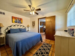 A bedroom with a bed, a ceiling fan, a painting, a lamp, a rug, and a view of the kitchen.