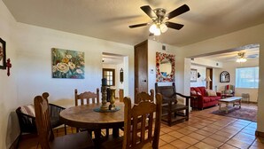 A dining area with a round table, wooden chairs, a ceiling fan, and a floral painting on the wall.