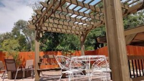 An outdoor patio with a wooden pergola, white metal chairs, and a table.