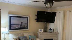 Een woonkamer met een beige bank, een houten salontafel, een open haard, een plafondventilator en een flatscreen tv aan de muur.