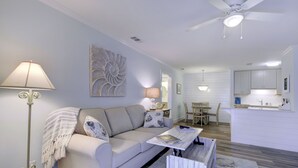 Ein modernes Wohnzimmer mit einer Couch, einem Couchtisch und einer Stehlampe. Es gibt einen Essbereich mit Tisch und Stühlen. Im Hintergrund ist eine Küche mit weißen Schränken und einem Deckenventilator zu sehen.