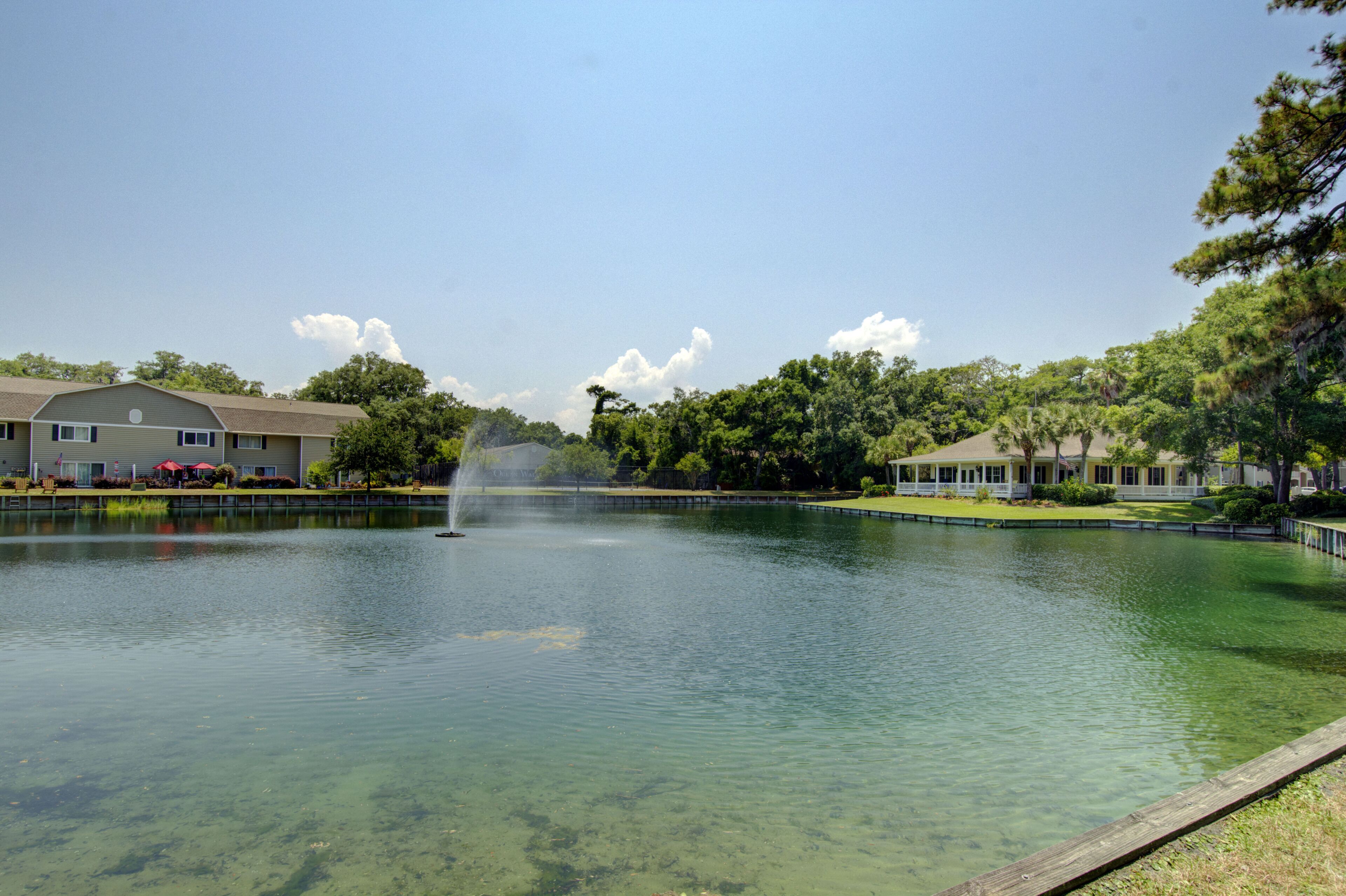 A clear lake with a fountain, surrounded by buildings with a white fence and trees.