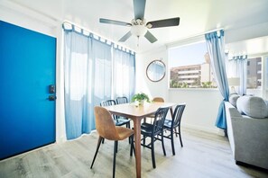 A room with a dining table, chairs, a ceiling fan, a sofa, and a view of buildings outside.