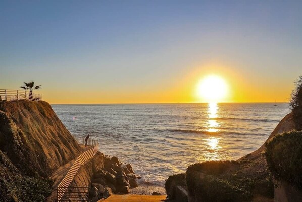 A coastal sunset view with a rocky shoreline, a staircase leading down to the water, and a person standing at the top.
