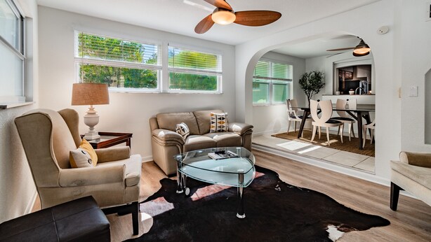 A modern living room with a glass coffee table, leather armchairs, a ceiling fan, and a dining area visible through an archway.