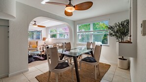 A dining area with a glass table, chairs, a ceiling fan, and a view of the living room.