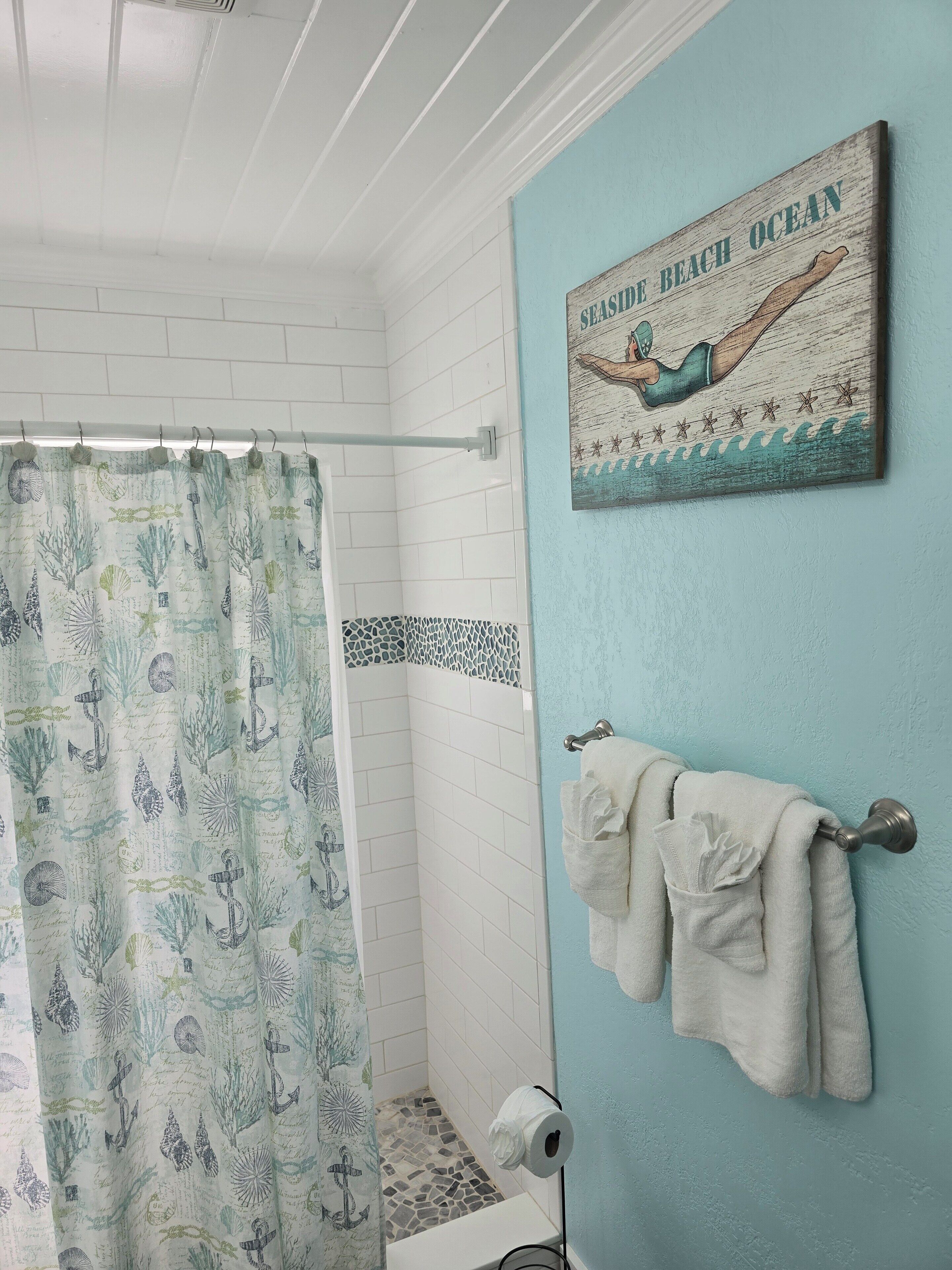 Une salle de bain avec un rideau de douche orné de motifs marins, un porte-serviettes avec des serviettes et une pancarte fixée au mur sur laquelle on peut lire : « Plage en bord de mer ».