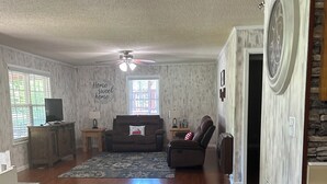A spacious living room with hardwood floors, a ceiling fan, a fireplace, and a large window. - Cedar Mountain Lodge (Bryant)