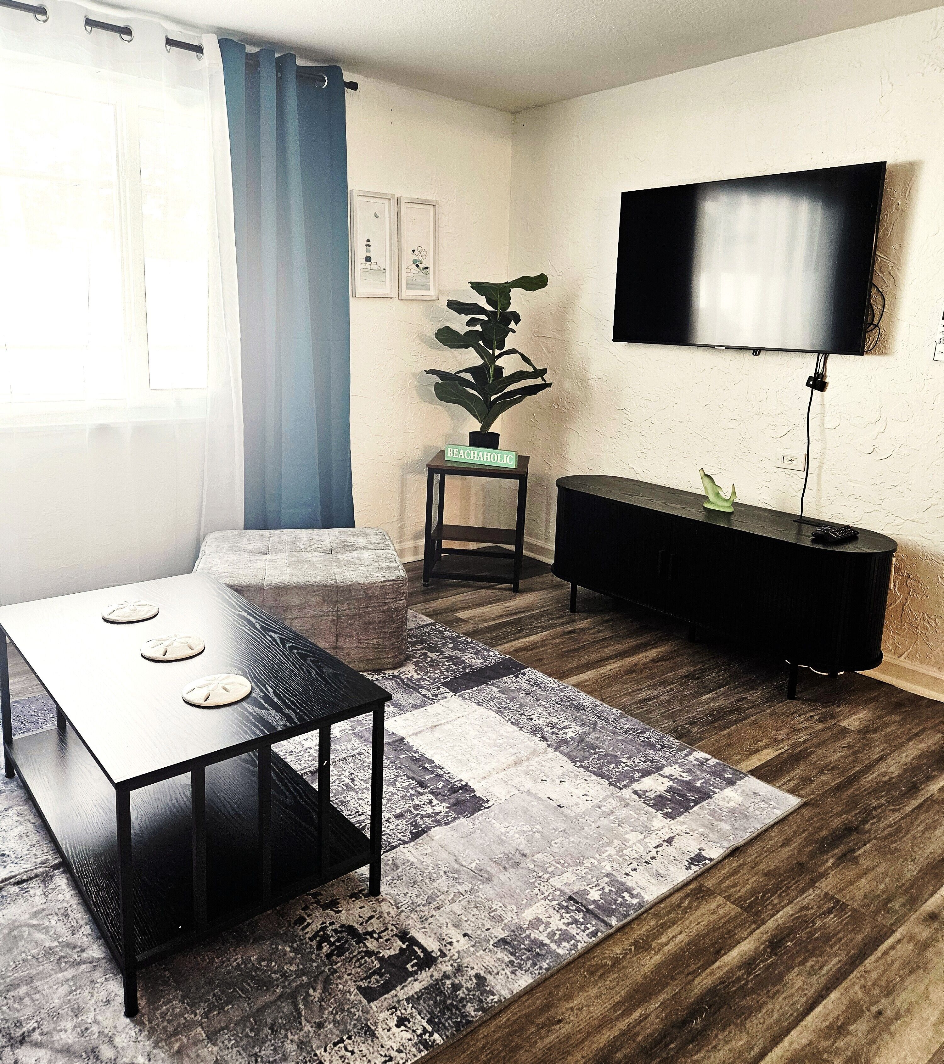 Ein modernes Wohnzimmer mit Holzboden, einem schwarzen Couchtisch, einem grauen Sofa, einem kleinen Beistelltisch mit Pflanze und einem flachbildfernseher an der Wand.