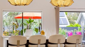 A dining area with four white chairs, a table with a potted plant and a vase, and a window with a view of greenery and a house. - KAMALA - MOUNT ELIZA (Mount Eliza)