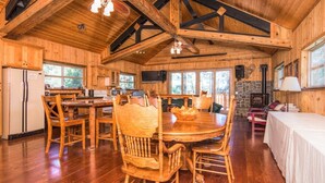 A spacious wooden cabin with a large dining table, wooden chairs, a stone fireplace, and a television mounted on the wall.