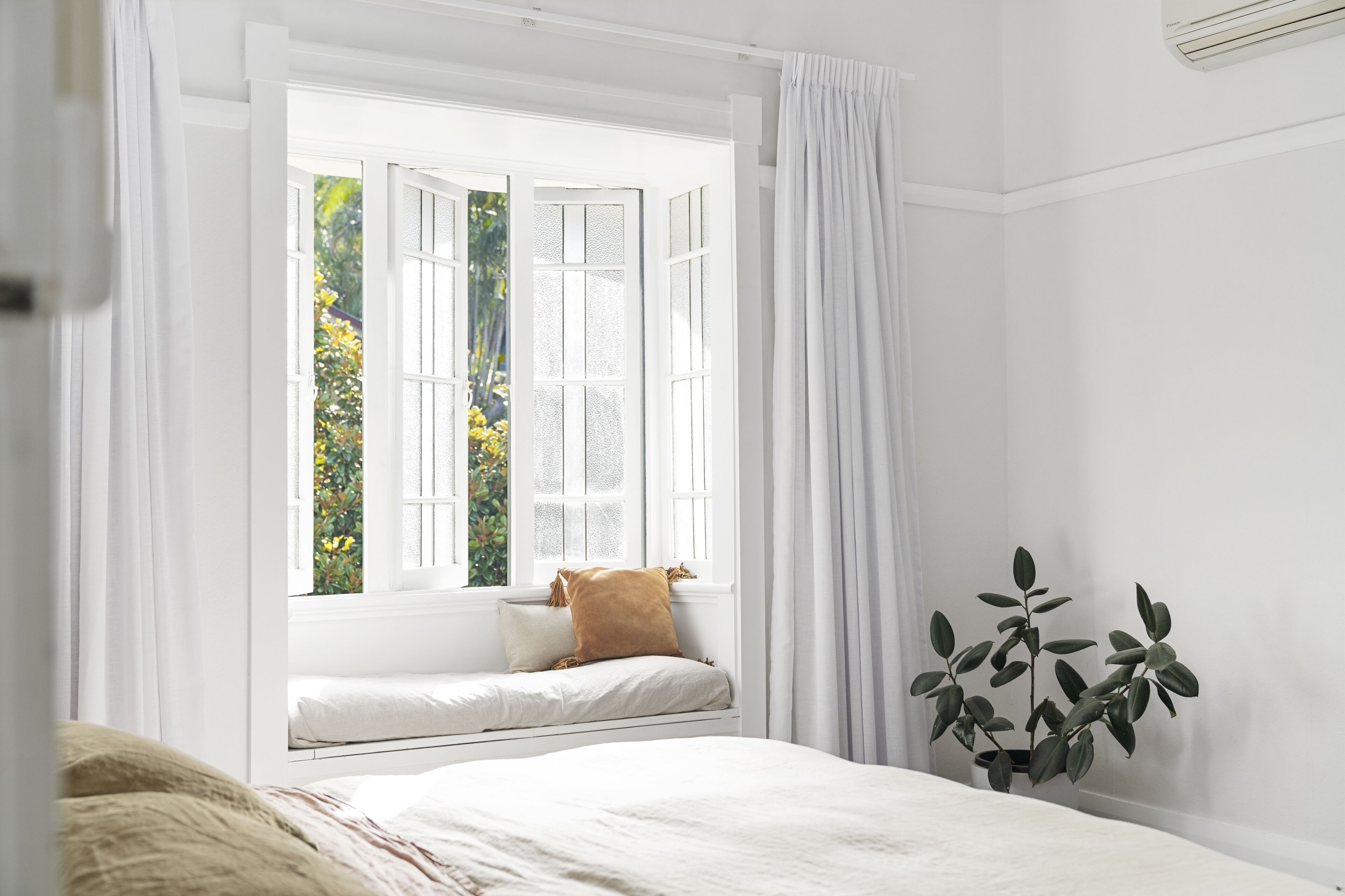 A bedroom with a large window, a bed, a bedside plant, and a window seat.
