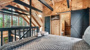 A wooden cabin bedroom with a bed, a view of the forest, and a sliding door to a bathroom. - Mountain Views|Hot Tub|Foosball|Minicade (Pigeon Forge)