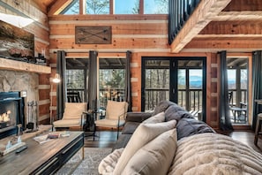 L’intérieur d’une cabane en rondins, avec une cheminée, un coin salon et de grandes fenêtres.