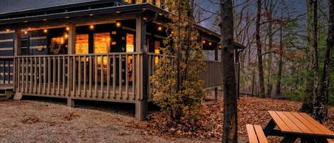 A cabin with a deck and a picnic table, surrounded by trees and a starry sky.