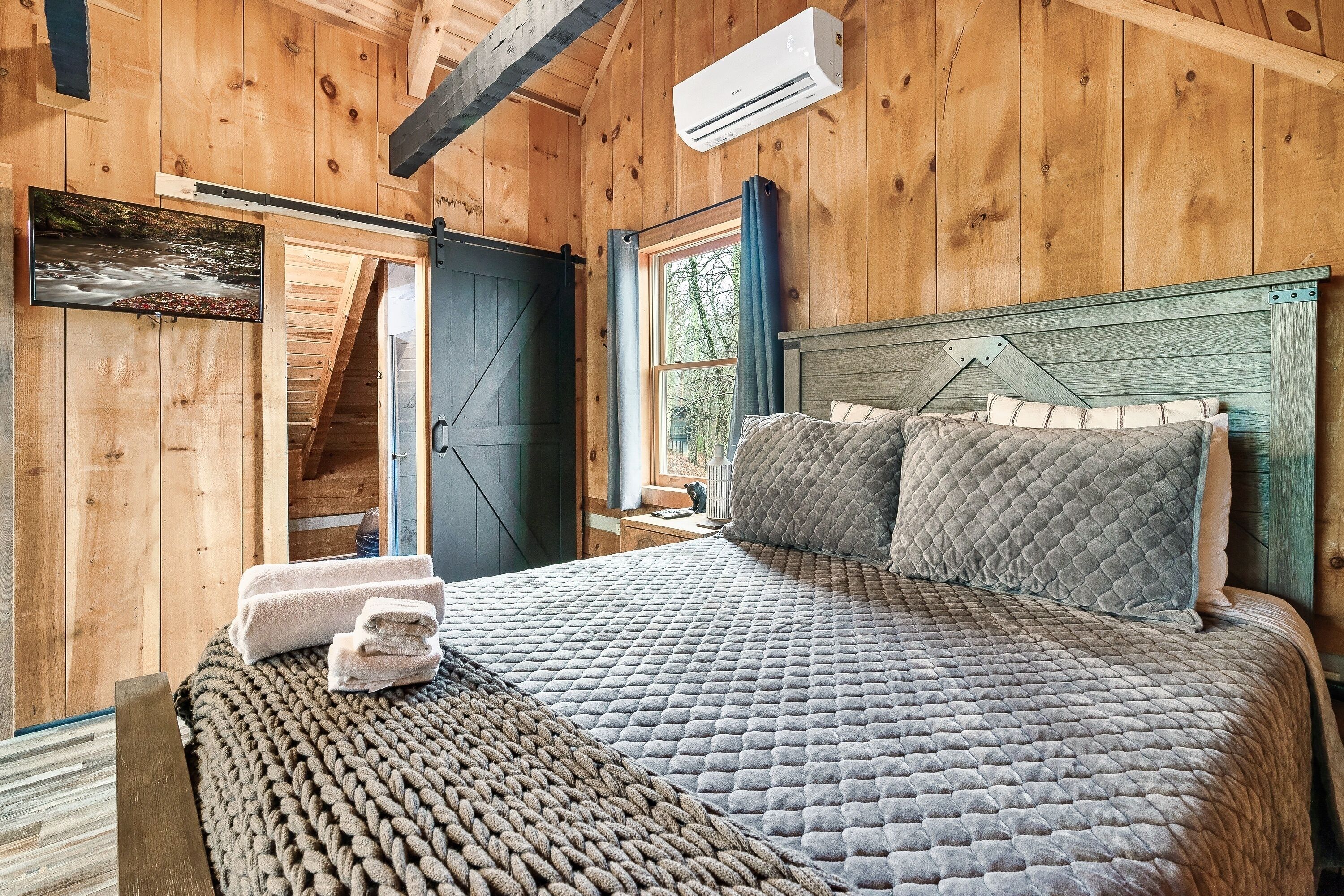 Dormitorio de cabaña de madera con cama, almohadas, una manta, un equipo de aire acondicionado y una ventana con cortinas.