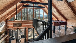 A spacious attic room with a wooden ceiling, a foosball table, and a large window offering a view of the surrounding forest. - Mountain Views|Hot Tub|Foosball|Minicade (Pigeon Forge)
