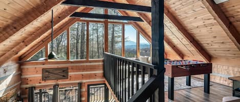 Ein geräumiges Dachzimmer mit Holzbalkendecke, einem Kicker und einem großen Fenster mit Blick auf den umliegenden Wald.
