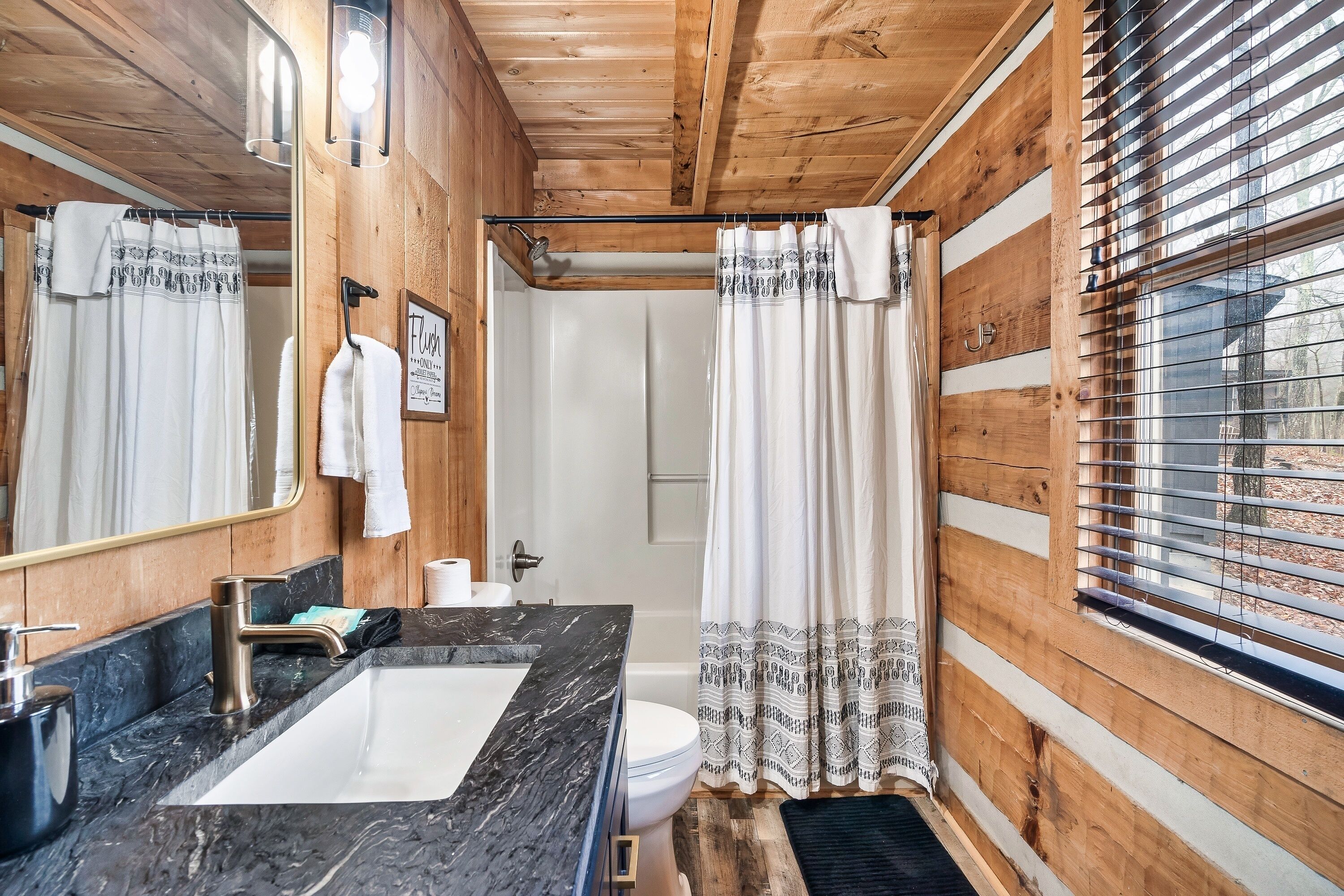 Baño de cabaña de madera con lavamanos de mármol, inodoro, ducha con cortina estampada y ventana con persianas.