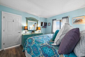 Une chambre avec une couvre-lit à motifs, une porte blanche, une commode turquoise, un miroir, un téléviseur et une fenêtre avec des rideaux.