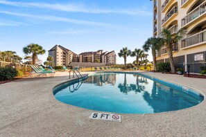 Une piscine avec un panneau indiquant une profondeur minimale de 1,5 mètre (5 pieds) et interdisant la plongée, entourée de palmiers et bordée d’un immeuble doté de balcons.