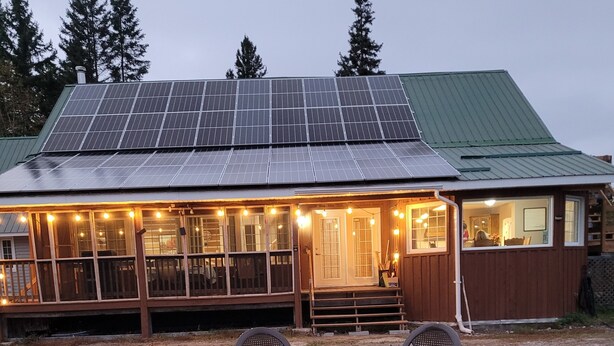 A rustic cabin with a solar panel roof, a covered porch with string lights, and a fire pit in the foreground. - SLOPES AND STREAMS (Searchmont)