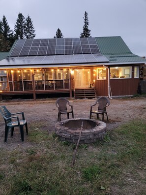 Une cabane rustique avec un toit équipé de panneaux solaires, une véranda couverte illuminée de guirlandes lumineuses et un foyer au premier plan.