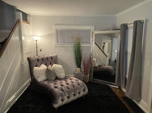 Une chambre à coucher avec un lit doté d’une tête de lit capitonnée, une table de chevet, une lampe, une plante en pot et un miroir.