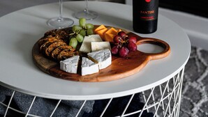A round white table with a wooden board holding cheese, grapes, and crackers, accompanied by two wine glasses and a bottle of wine.